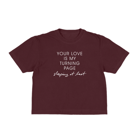 A maroon short-sleeve t-shirt with white printed text on the front reading 'YOUR LOVE IS MY TURNING PAGE sleeping at last'